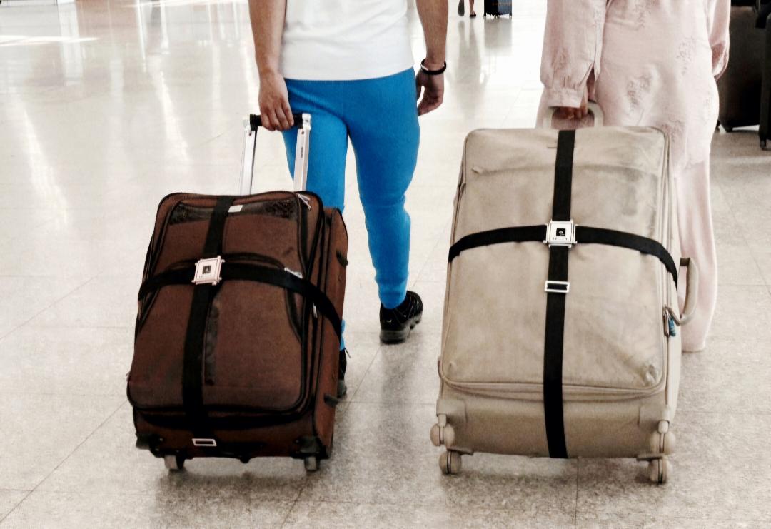 Two people with suitcases at an airport, one in blue pants and a white shirt, the other in a light-colored outfit.